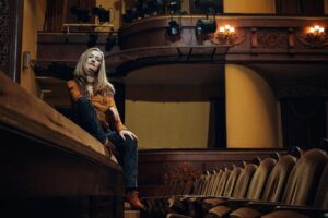 Une femme blonde, vêtue d’une blouse orange et d’un pantalon bleu, observe un théâtre vide depuis une loge, dans une ambiance théâtrale chaleureuse.